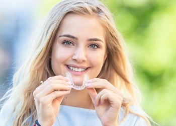 Smiling blonde woman holding an Invisalign tray near her mouth